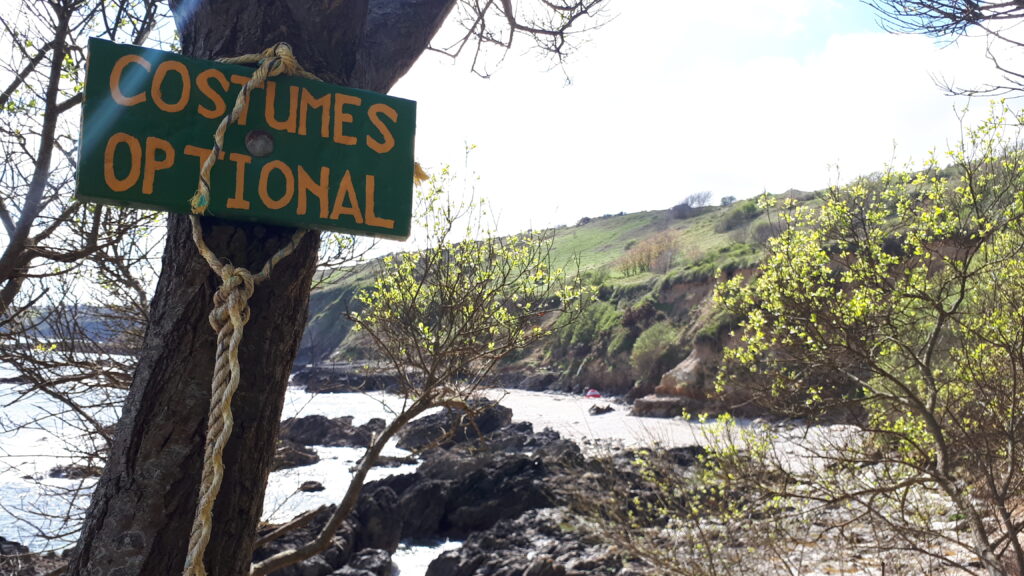 sign pinned to a tree above a rocky beach reading 'costumes optional'